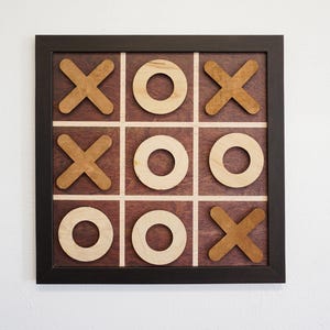 May include: A square wooden tic-tac-toe game board featuring a dark brown frame and a dark stained wood grid. The game includes light-colored wooden circles and medium-brown wooden X-shaped pieces arranged in a three-by-three grid pattern.