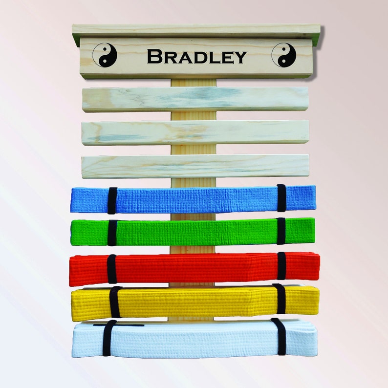 K&ouml;nnte beinhalten: Ein h&ouml;lzernes Display f&uuml;r Kampfsportg&uuml;rtel mit dem Namen "BRADLEY" oben aufgedruckt. Das Display h&auml;lt mehrere farbige G&uuml;rtel: blau, gr&uuml;n, rot, gelb und wei&szlig;. Yin-und-Yang-Symbole befinden sich auf beiden Seiten des Namens.