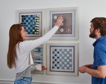 Op de afbeelding: Twee mensen spelen spelletjes op een muur met vier ingelijste spelborden. De borden zijn allemaal verschillende spellen, waaronder dammen, tic-tac-toe en een spel met schelpen. De borden zijn allemaal in een lichtblauw en wit kleurenschema.