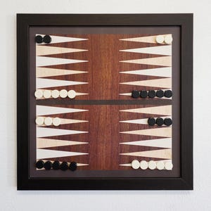 May include: A square backgammon board mounted in a black frame. The board features a dark brown wood grain center panel with alternating light cream and dark brown triangular points. Black and white circular game pieces are arranged on the points.
