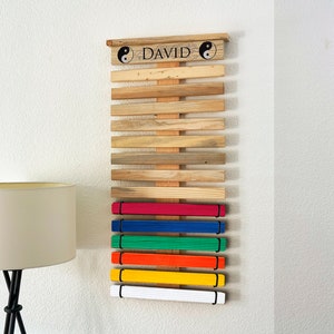 K&ouml;nnte beinhalten: Holz-G&uuml;rtel-Display f&uuml;r Kampfsportarten mit dem Namen "DAVID" und Yin-Yang-Symbolen. Es zeigt G&uuml;rtel in verschiedenen Farben: Wei&szlig;, Gelb, Orange, Gr&uuml;n, Blau und Rot. Das Display ist an einer wei&szlig;en Wand befestigt.