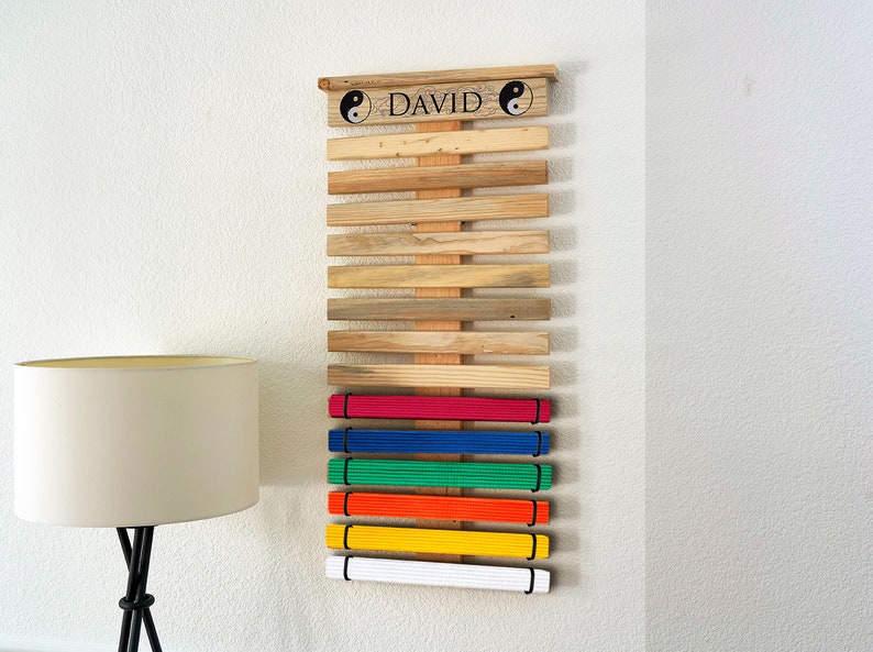 K&ouml;nnte beinhalten: Holz-G&uuml;rtel-Display f&uuml;r Kampfsportarten mit dem Namen "DAVID" und Yin-Yang-Symbolen. Es zeigt G&uuml;rtel in verschiedenen Farben: Wei&szlig;, Gelb, Orange, Gr&uuml;n, Blau und Rot. Das Display ist an einer wei&szlig;en Wand befestigt.