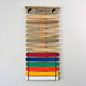 K&ouml;nnte beinhalten: Holz-G&uuml;rtel-Display f&uuml;r Kampfsportarten mit dem Namen "DAVID" und Yin-Yang-Symbolen. Es enth&auml;lt farbige G&uuml;rtel: rot, blau, gr&uuml;n, orange, gelb und wei&szlig;. Die Holzlatten haben verschiedene Braun- und Graut&ouml;ne.