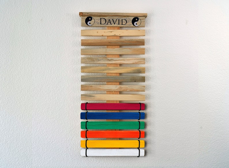 K&ouml;nnte beinhalten: Holz-G&uuml;rtel-Display f&uuml;r Kampfsportarten mit dem Namen "DAVID" und Yin-Yang-Symbolen. Es enth&auml;lt farbige G&uuml;rtel: rot, blau, gr&uuml;n, orange, gelb und wei&szlig;. Die Holzlatten haben verschiedene Braun- und Graut&ouml;ne.