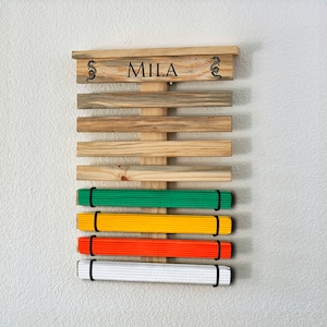 K&ouml;nnte beinhalten: Holz-G&uuml;rtel-Display f&uuml;r Kampfsportarten mit dem Namen "MILA" oben. Das Display hat horizontale Holzlatten und h&auml;lt gr&uuml;ne, gelbe, orangefarbene und wei&szlig;e G&uuml;rtel. Drachendesigns flankieren den Namen.