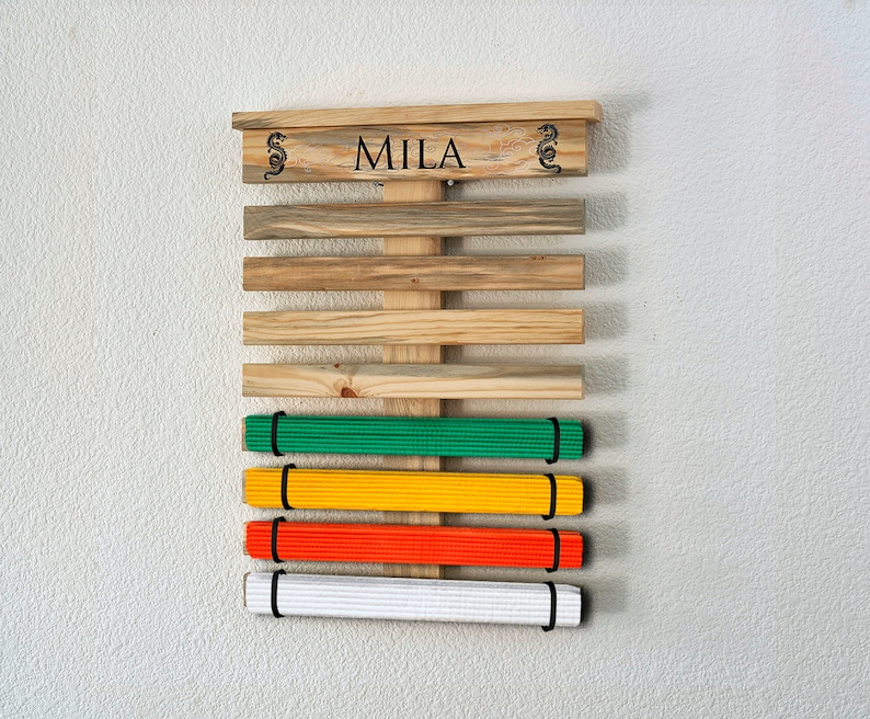 K&ouml;nnte beinhalten: Holz-G&uuml;rtel-Display f&uuml;r Kampfsportarten mit dem Namen "MILA" oben. Das Display hat horizontale Holzlatten und h&auml;lt gr&uuml;ne, gelbe, orangefarbene und wei&szlig;e G&uuml;rtel. Drachendesigns flankieren den Namen.
