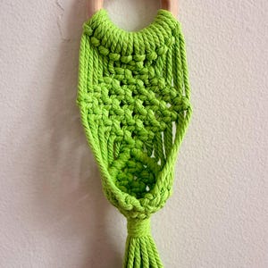 May include: A lime green macrame wall hanging featuring a circular light wood ring at the top. The cord is knotted in a complex geometric pattern that tapers downward, ending in a thick, straight fringe tassel at the bottom of the piece.