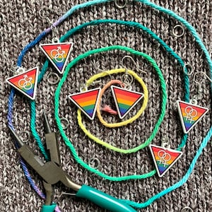 May include: A set of rainbow triangle earrings with gender symbols. The earrings are on a background of colorful yarn.