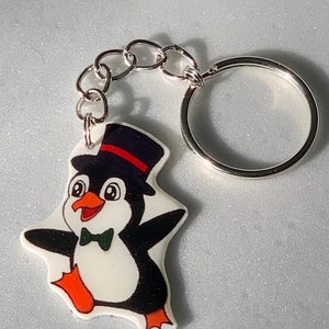 May include: A white penguin wearing a black top hat and a green bow tie. The penguin is holding its arms out and has a happy expression on its face. The penguin is attached to a silver key chain.