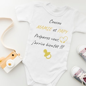 Puede incluir: Un body blanco para bebé con el texto "Coucou Mamie et Papy Preparez vous, j'arrive bientot!!!" en negro. Un corazón dorado con una flecha está por encima del texto. Un chupete dorado está debajo del texto. El body está sobre un fondo blanco con una cámara de madera y dos zapatos de bebé grises.