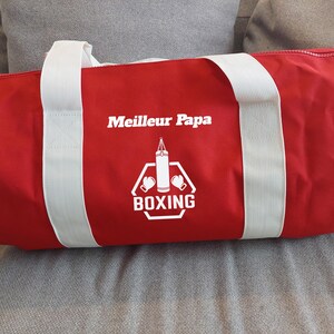 May include: Red canvas duffel bag with white straps and a white graphic of a boxing bag and gloves with the text "Meilleur Papa BOXING".