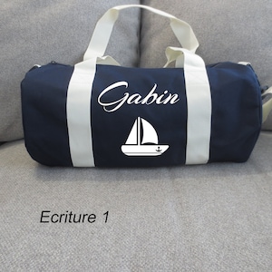 Puede incluir: Una bolsa de lona azul marino con correas blancas y un gráfico de velero blanco. La bolsa está personalizada con el nombre "Gabin" en letras blancas cursivas.