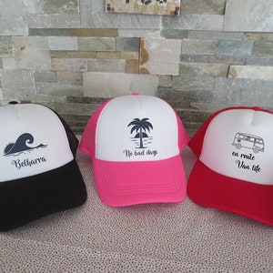 Peut inclure: Trois casquettes de camionneur avec des designs différents. La première casquette est noire avec une vague blanche et le texte "Belharra". La deuxième casquette est rose avec un palmier blanc et le texte "No bad days". La troisième casquette est rouge avec un van blanc et le texte "en route Van life".