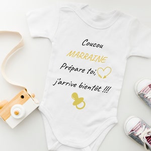 Peut inclure: Combinaison blanche pour bébé avec le texte "Coucou Marraine Prépare toi j'arrive bientôt!!!" en noir. La combinaison a un cœur et une flèche dorés ainsi qu'une sucette dorée.