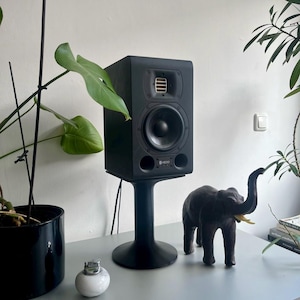 Könnte beinhalten: Ein schwarzer Studio-Monitor-Lautsprecher auf einem schwarzen Ständer mit einer kleinen Elefantenfigur auf einer hellgrauen Oberfläche.