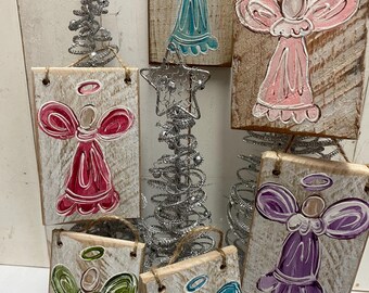 May include: A collection of handmade wooden angel ornaments in various colors, including pink, teal, green, and purple. Each ornament features a painted angel design with a halo and is attached to a rustic wooden plank with twine for hanging. Decorative silver wire Christmas tree and star accents are also visible.