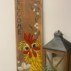 May include: Wooden sign with the word "WELCOME" painted vertically in black. A yellow chicken with red wattle and comb is painted on the sign, looking at the viewer. The chicken is surrounded by green vines with white flowers.