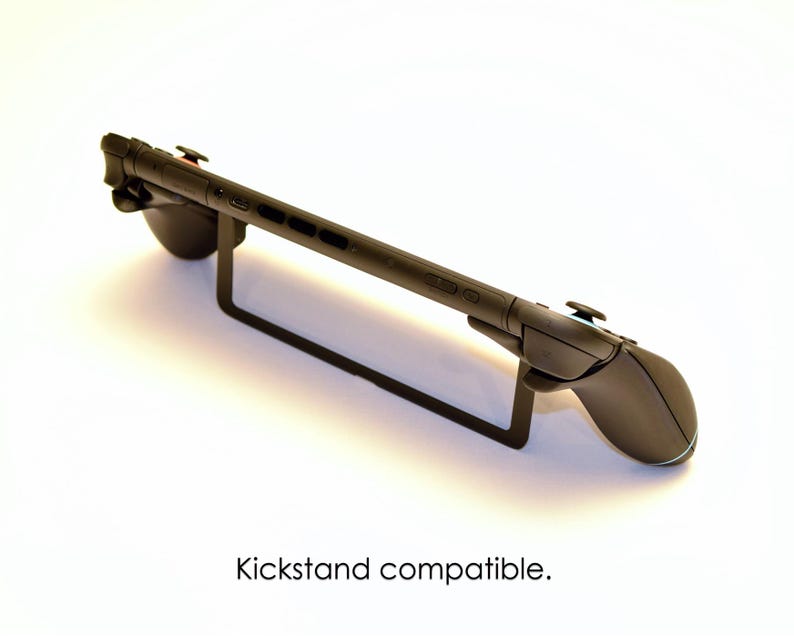 May include: A black gaming console with attached controllers is displayed on a black metal stand. The text "Kickstand compatible" is at the bottom of the image. The console is designed for handheld gaming.