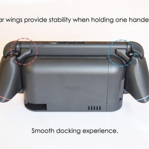 May include: A black gaming console accessory with attached side grips. The text "Rear wings provide stability when holding one handed" and "Smooth docking experience" are visible. The accessory has a Nintendo logo.