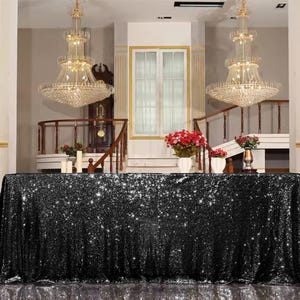 Könnte beinhalten: Schwarzer Sequinentischdecke, die über einen Tisch in einem Raum mit zwei großen Kristallleuchtern drapiert ist. Die Tischdecke ist ein funkelnder, luxuriöser Stoff.