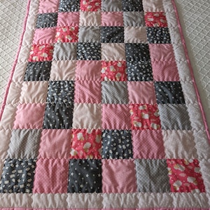 May include: A pink and grey patchwork quilt with a variety of patterns, including stars, dots, and sheep. The quilt is edged with a pink border.