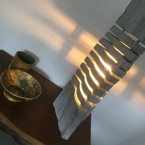 Può includere: Una lampada in legno con un design unico, composta da pezzi di legno impilati con tagli che permettono alla luce di passare attraverso. La lampada è accesa e proietta un bagliore caldo.