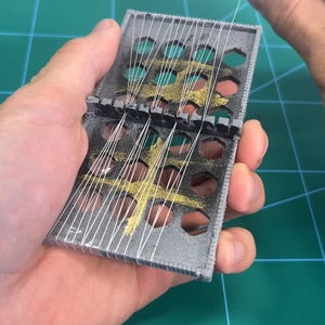 May include: A gray, hinged, rectangular object with multiple holes and gold accents, strung with white threads. The object is held in a person's hand against a green surface.