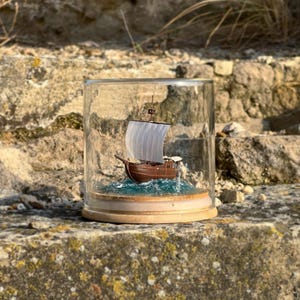 May include: A miniature wooden ship with white sails is displayed in a glass jar on a wooden base. The ship is sailing on a blue and green sea with white foam waves. The jar is sitting on a stone surface.