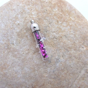 May include: A small, clear glass vial pendant filled with bright pink glitter. The vial has a silver-colored metal cap and a small loop for hanging. The pendant is held by a clear, curved plastic holder.