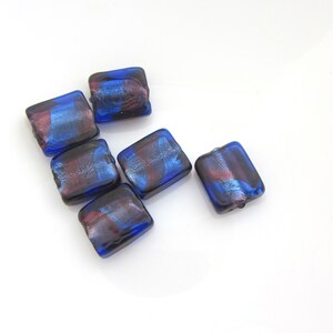 Peut inclure: Six perles carrées en verre aux bords arrondis. Les perles sont d'un bleu profond avec des stries rouges et argentées. Les perles sont disposées sur une surface blanche, probablement pour la fabrication de bijoux.