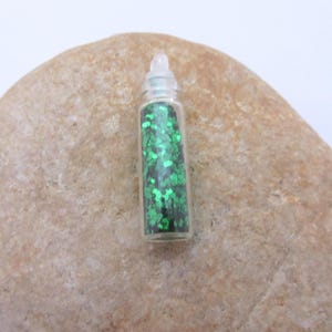 May include: A small clear glass bottle filled with green glitter. The glitter has a leaf-like shape. The bottle has a clear plastic stopper. The bottle is resting on a light brown stone.