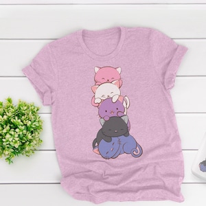 May include: A pale pink t-shirt with a cartoon design of stacked cats in pink, white, purple, and black. The cats are smiling. A matching mug with the same design is also visible.