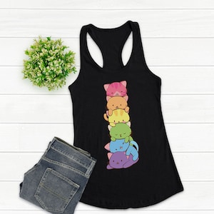 May include: Black racerback tank top featuring a stack of six colorful cartoon cats. The cats are in shades of pink, orange, yellow, green, blue, and purple. The tank top is paired with blue jeans and a small green plant.