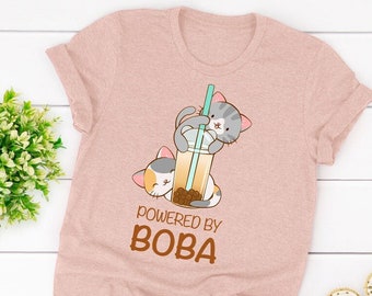 May include: A pale pink t-shirt with a cartoon illustration of two cats and a boba drink. The text "POWERED BY BOBA" is printed in brown. The shirt is made of a soft material and is suitable for casual wear.