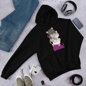 Puede incluir: Sudadera con capucha negra con una pila de cuatro gatos de dibujos animados en diferentes colores, incluyendo gris, blanco y morado. Los gatos est&aacute;n apilados uno encima del otro, con el gato m&aacute;s grande en la parte inferior y el gato m&aacute;s peque&ntilde;o en la parte superior.