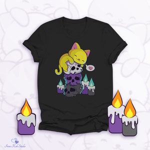 Puede incluir: Camiseta negra con un gato amarillo durmiendo sobre una pila de calaveras. El diseño incluye velas moradas con llamas azules y una burbuja de diálogo con un corazón. La camiseta tiene cuello redondo.