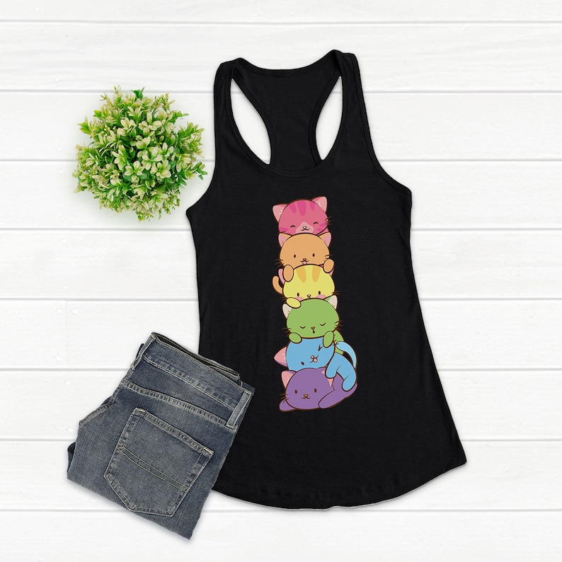May include: Black racerback tank top featuring a stack of six colorful cartoon cats. The cats are in shades of pink, orange, yellow, green, blue, and purple. The tank top is paired with blue jeans and a small green plant.