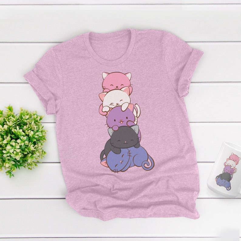 Pu&ograve; includere: T-shirt rosa chiaro con un disegno a cartoni animati di gatti impilati in rosa, bianco, viola e nero. I gatti sorridono. &Egrave; visibile anche una tazza abbinata con lo stesso disegno.