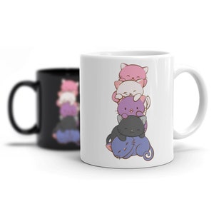 May include: White ceramic mug with a design of stacked cartoon cats in pink, white, purple, and black. The cats are smiling and stacked. A blurred black mug is visible in the background.