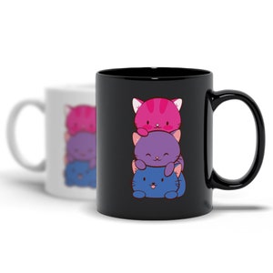May include: Black ceramic mug featuring a stack of three cartoon cats in pink, purple, and blue, with the colors representing the bisexual pride flag. A second white mug with a similar design is slightly out of focus in the background.