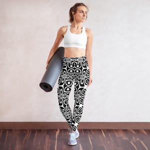 Könnte beinhalten: Schwarz-weiß gemusterte Leggings mit passendem Sport-BH. Die Leggings haben ein sich wiederholendes geometrisches Design. Eine zusammengerollte graue Yogamatte wird in der linken Hand gehalten. Die Person trägt graue Sportschuhe.