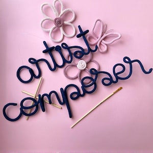 Op de afbeelding: Roze achtergrond met de woorden "artist composer" in marineblauw touw. Decoratieve bloemen en een vlinder van touw zijn ook aanwezig. Een penseel en kleine houten stokjes maken het artistieke thema compleet.