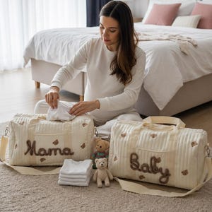 Könnte beinhalten: Cremefarbene, gesteppte Wickeltaschen mit den braunen Schriftzügen "Mama" und "Baby". Die Taschen haben Teddybär-Akzente und werden von kleinen Teddybären und gefalteten weißen Bodys begleitet. Die Szene spielt in einem Schlafzimmer.
