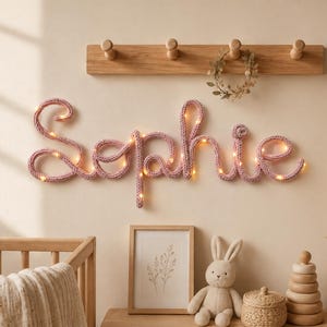 Può includere: Una scritta da parete con il nome Sophie in filato lavorato a maglia rosa, illuminata da luci a LED bianco caldo integrate. Le lettere in corsivo sono montate orizzontalmente, caratterizzate da una consistenza morbida e un effetto luminoso.
