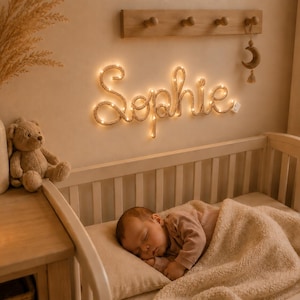 Könnte beinhalten: Ein an der Wand montiertes LED-Lichtschild, das den Namen Sophie in Schreibschrift anzeigt und warmweiß leuchtet. Das Schild befindet sich über einem Holzbettchen, in dem ein Baby schläft, ergänzt durch eine Holzhakenleiste und einen Mondanhänger.