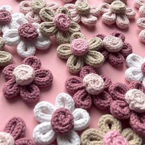 Op de afbeelding: Een verzameling handgemaakte gehaakte bloemen in verschillende kleuren, waaronder wit, roze en beige. De bloemen zijn gerangschikt op een roze oppervlak, waarbij hun gedetailleerde bloemblaadjesontwerp en getextureerde centra worden getoond. Ideaal voor knutselprojecten.