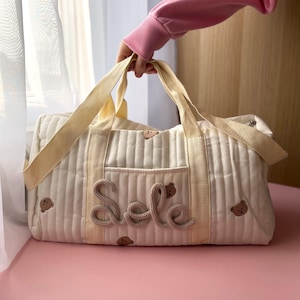 Puede incluir: Bolso tote acolchado de color crema con asas y ribetes beige. La bolsa presenta el nombre "Sole" en una fuente cursiva y pequeñas aplicaciones de osos. La bolsa es sostenida por una persona que lleva un suéter rosa.