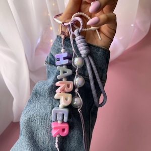 Può includere: Un portachiavi personalizzato con il nome "HARPER" scritto con perline colorate in stile blocco. Il portachiavi presenta un cordino lavanda, perline iridescenti e una chiusura color argento. Lo sfondo include una giacca di jeans e uno sfondo rosa.