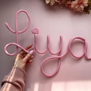 Puede incluir: El nombre "Liya" en rosa, tejido a ganchillo, se muestra sobre un fondo rosa. Las letras están hechas de un hilo grueso y suave, con una mano que sostiene la "L". Decoraciones florales son visibles en la parte superior. El nombre es un artículo decorativo.