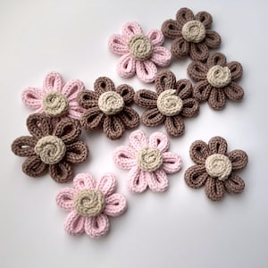 Puede incluir: Una colección de diez flores de ganchillo en rosa, marrón y beige. Las flores tienen un diseño sencillo con cinco pétalos y un círculo central.